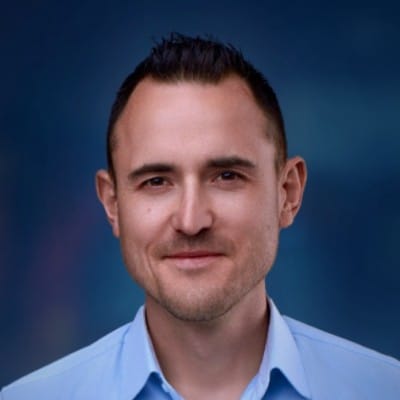 a man with short dark hair wearing a light blue collared shirt against a blurred blue background smiling confidently at the camera digital production A man with short dark hair wearing a light blue collared shirt against a blurred blue background, smiling confidently at the camera.