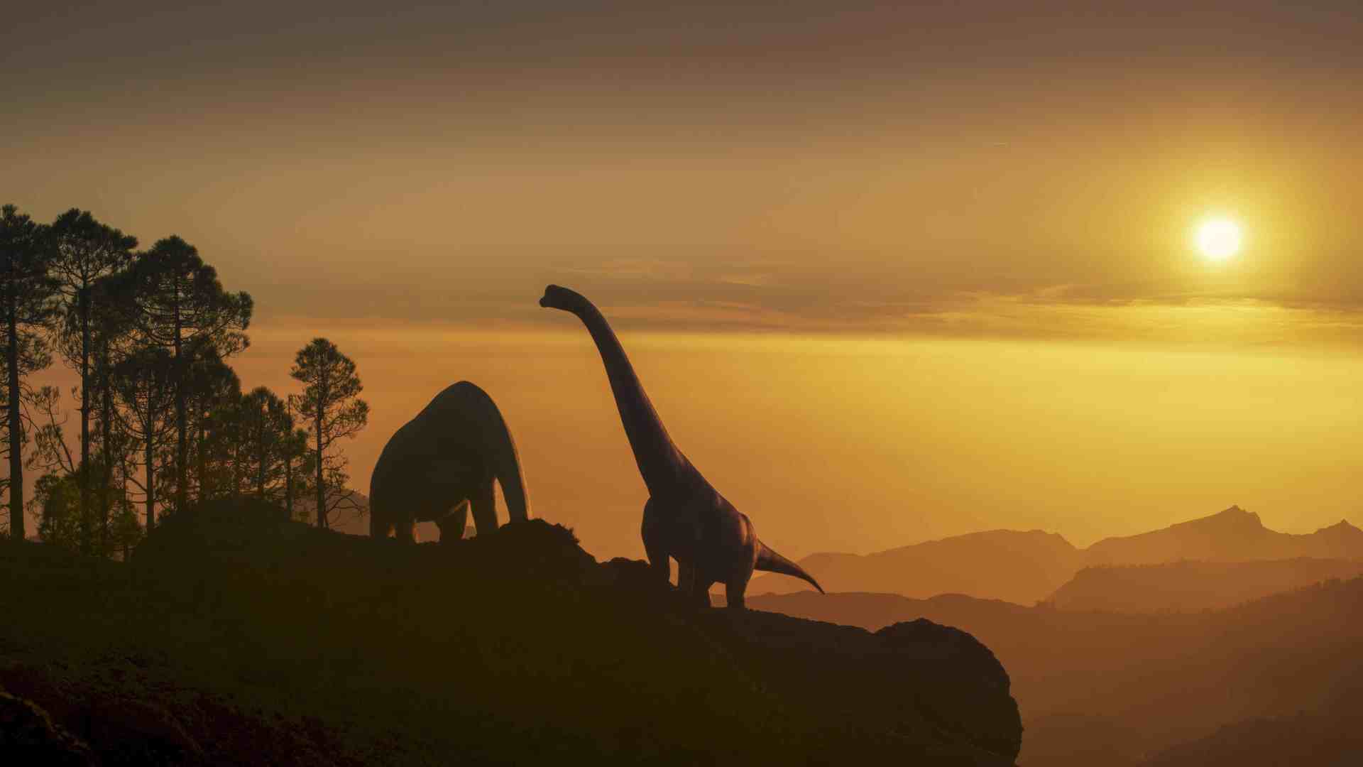 two towering dinosaurs silhouetted against a vibrant sunset standing on a rocky terrain with distant hills and trees in the background the sky is a blend of orange and yellow hues as the sun dips below the horizon digital production Two towering dinosaurs silhouetted against a vibrant sunset, standing on a rocky terrain with distant hills and trees in the background. The sky is a blend of orange and yellow hues as the sun dips below the horizon.