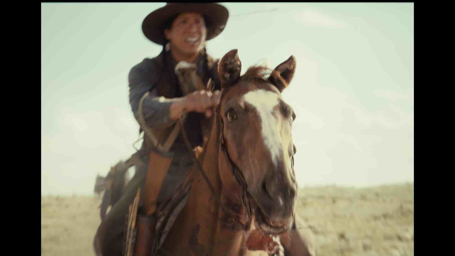 a person riding a brown horse on an open landscape wearing a widebrimmed hat and a denim outfit the rider appears to be smiling while holding the reins with a blurred background of dry terrain and blue sky digital production A person riding a brown horse on an open landscape, wearing a wide-brimmed hat and a denim outfit. The rider appears to be smiling while holding the reins, with a blurred background of dry terrain and blue sky.