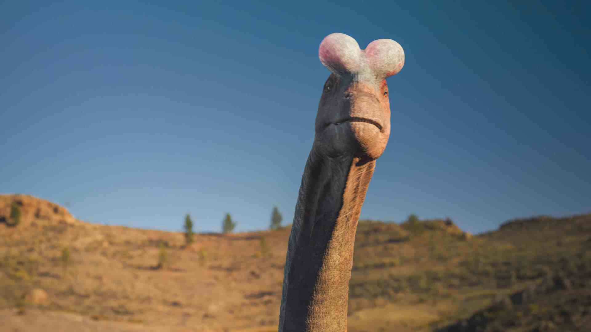 a whimsical dinosaur with a long neck and colorful rounded head features stands in a desert landscape against a clear blue sky the background reveals rocky terrain with sparse vegetation digital production A whimsical dinosaur with a long neck and colorful, rounded head features stands in a desert landscape against a clear blue sky. The background reveals rocky terrain with sparse vegetation.
