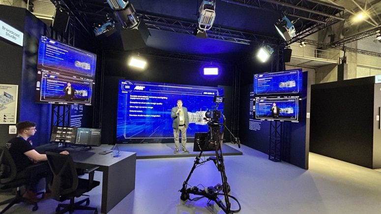 a modern studio setup featuring multiple screens displaying digital content a presenter in a grey suit stands in front of a large screen while a camera setup is positioned to capture the scene a technician sits at a workstation in the background digital production A modern studio setup featuring multiple screens displaying digital content. A presenter in a grey suit stands in front of a large screen, while a camera setup is positioned to capture the scene. A technician sits at a workstation in the background.