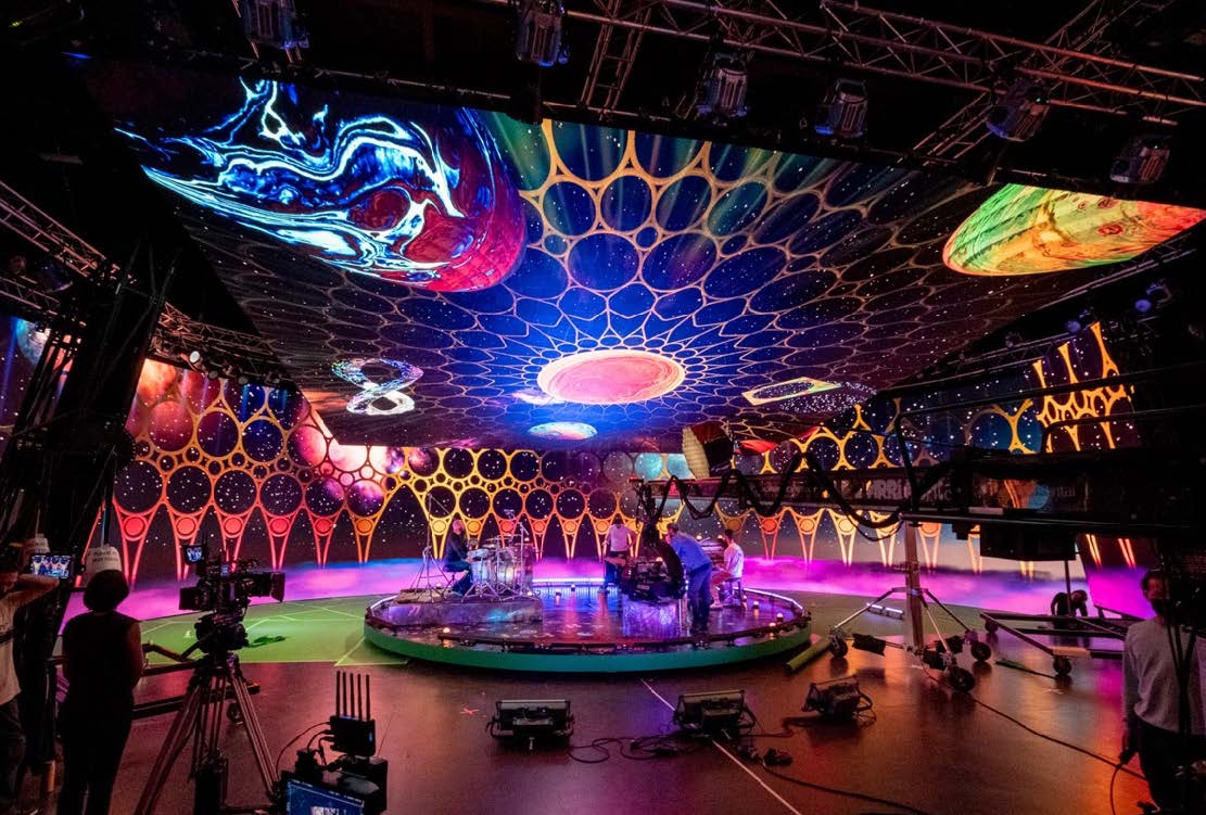 an elaborate stage setup featuring a circular platform surrounded by colorful cosmicthemed decorations and vibrant lights cameras and crew members are positioned around the stage capturing the dynamic atmosphere of the performance digital production An elaborate stage setup featuring a circular platform surrounded by colorful, cosmic-themed decorations and vibrant lights. Cameras and crew members are positioned around the stage, capturing the dynamic atmosphere of the performance.