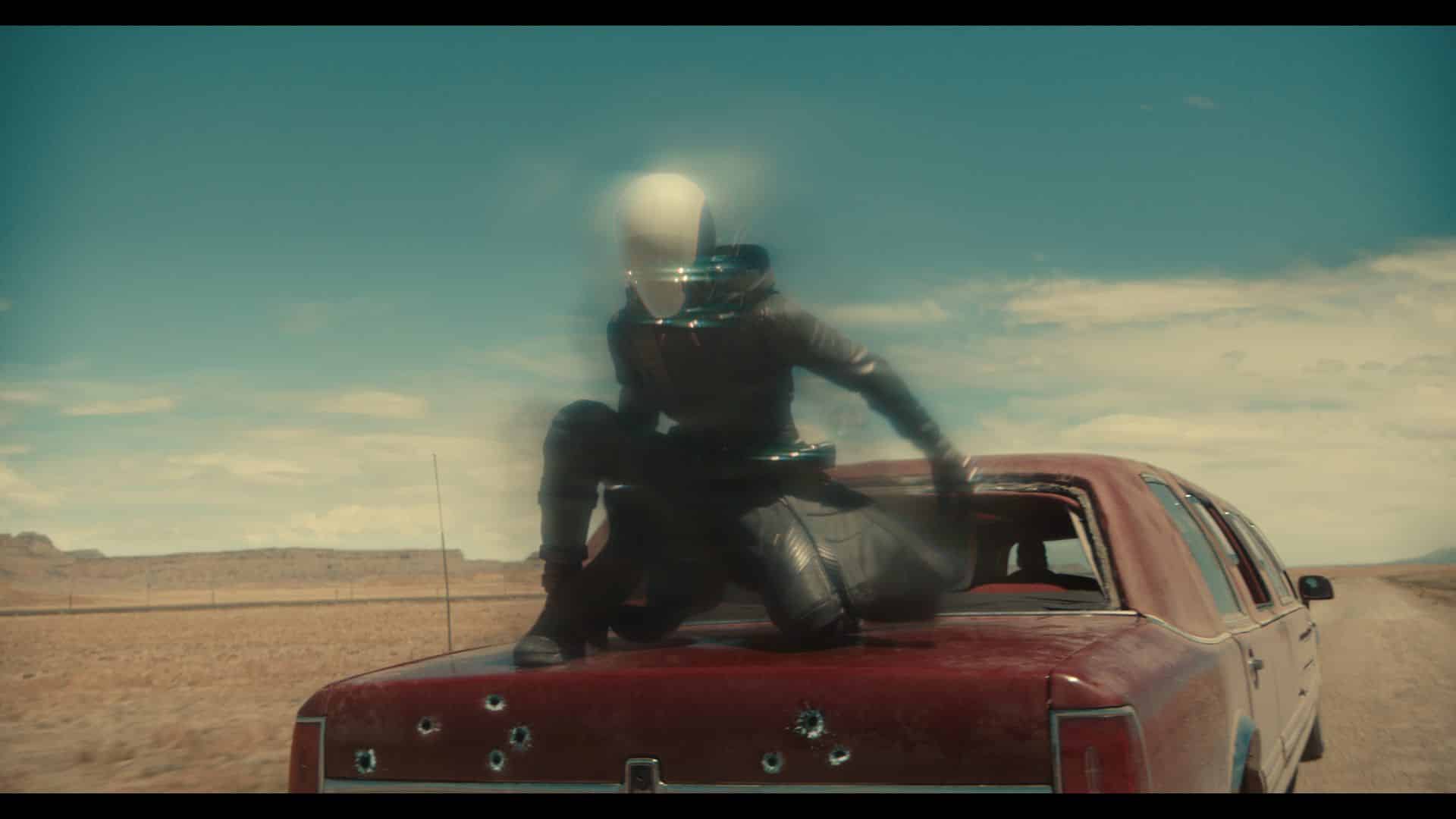 a figure in a dark outfit and pale helmet crouching on the roof of a weathered red car with visible bullet holes set against a vast desert landscape under a bright blue sky digital production A figure in a dark outfit and pale helmet crouching on the roof of a weathered red car with visible bullet holes, set against a vast desert landscape under a bright blue sky.