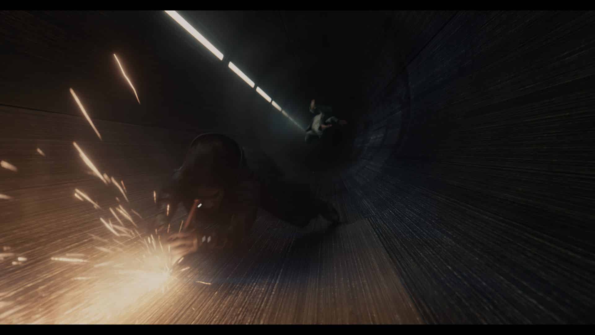 A dark tunnel with a figure sliding on the ground, causing sparks to fly. Another figure is seen in the background, adding depth to the scene. The tunnel walls are metallic with a streamlined texture, and the lighting creates a dramatic atmosphere.