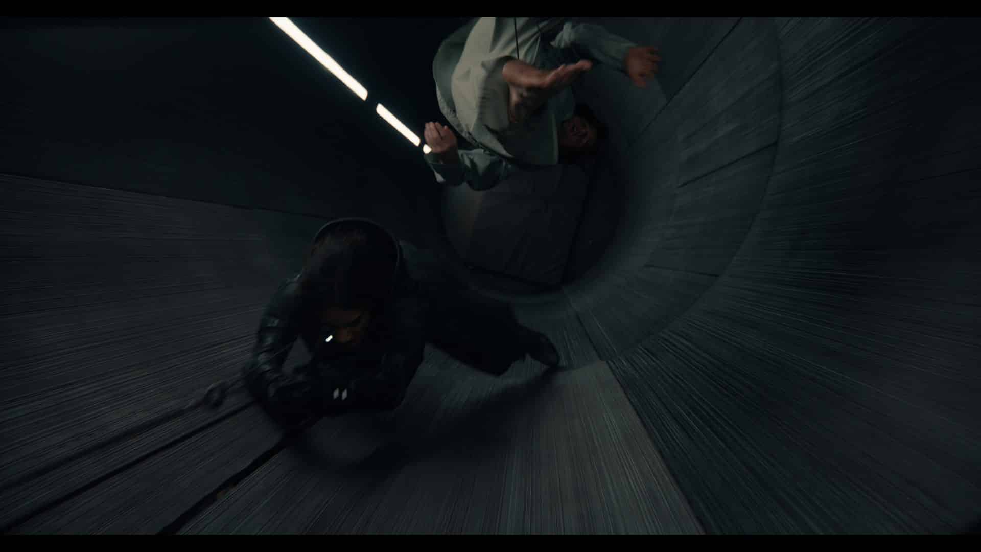 A dramatic action scene showing two individuals in a dark, cylindrical tunnel. One person is in a crouched position, pulling themselves forward, while the other appears to be falling backwards, their arms outstretched. Bright overhead lights illuminate the tunnel's surface, enhancing the sense of motion.