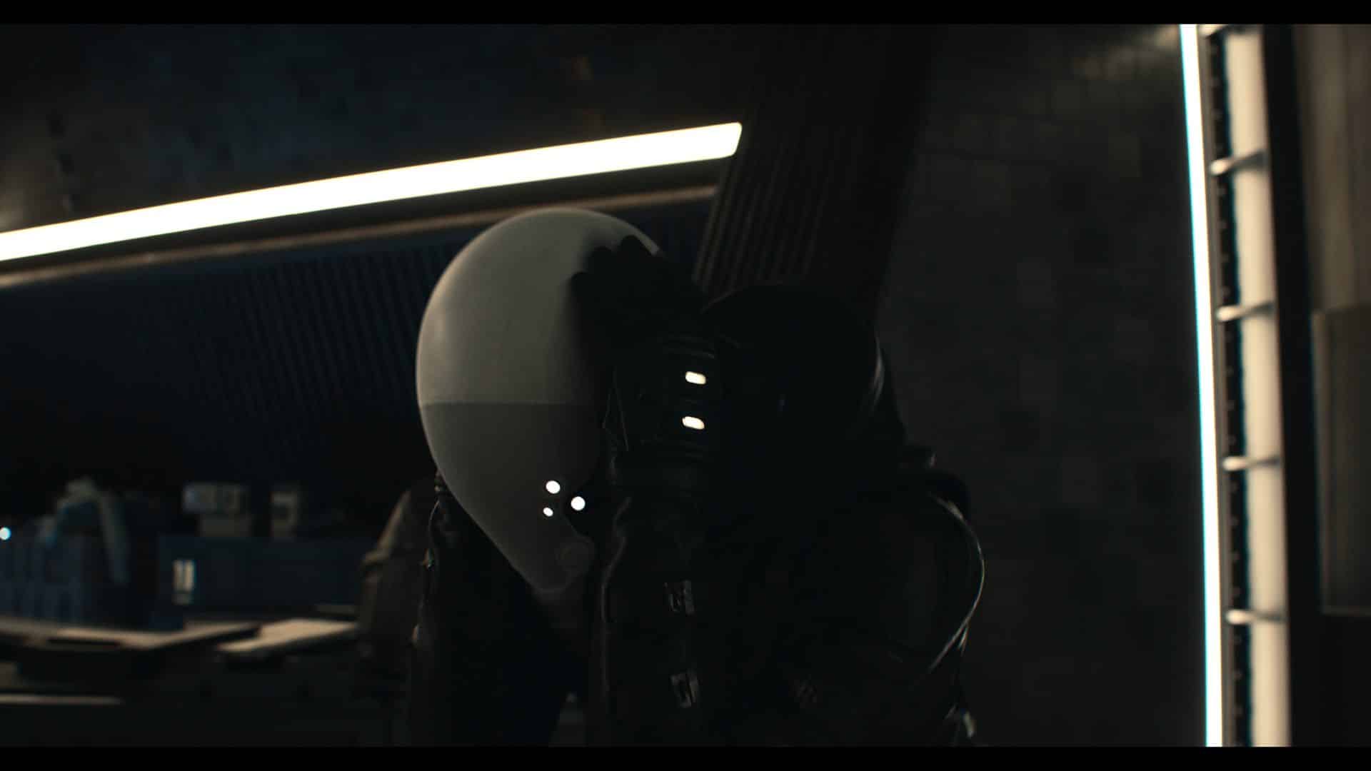 A humanoid figure with a smooth, white helmet and a black, armored suit stands in a dimly lit industrial setting, holding a large object. The background features modern architectural lines and storage units lit by neon lights.