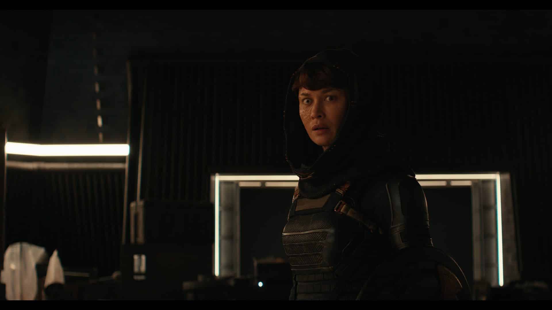 a person in a dark hightech environment wearing a hooded garment and a formfitting suit looking directly at the camera with a serious expression dim lighting highlights the contours of the scene with subtle reflections in the background digital production A person in a dark, high-tech environment wearing a hooded garment and a form-fitting suit, looking directly at the camera with a serious expression. Dim lighting highlights the contours of the scene, with subtle reflections in the background.