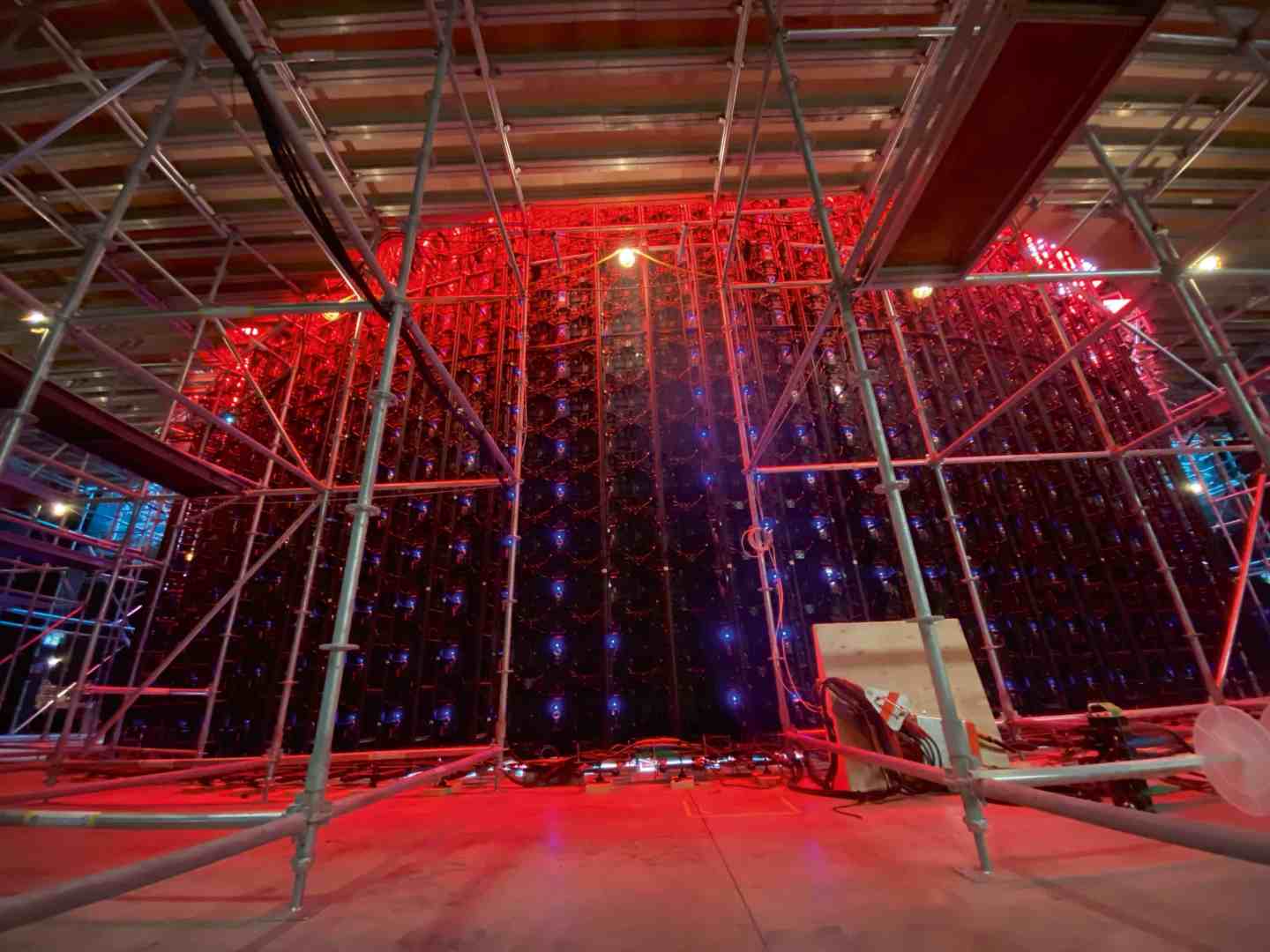 a construction site featuring scaffolding supporting a large led screen illuminated with red and blue lights the view is taken from the ground looking up at the vibrant display and metal framework against a dimly lit backdrop digital production A construction site featuring scaffolding supporting a large LED screen, illuminated with red and blue lights. The view is taken from the ground, looking up at the vibrant display and metal framework against a dimly lit backdrop.