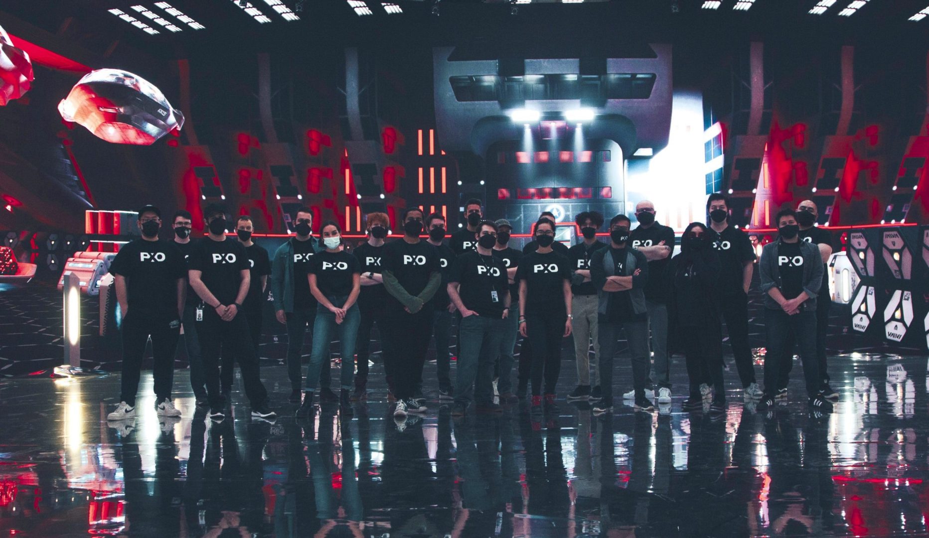 a diverse group of individuals standing in a semicircle on a glossy stage wearing matching black shirts with logos in front of a futuristic illuminated background featuring red and white elements digital production A diverse group of individuals standing in a semi-circle on a glossy stage, wearing matching black shirts with logos, in front of a futuristic, illuminated background featuring red and white elements.