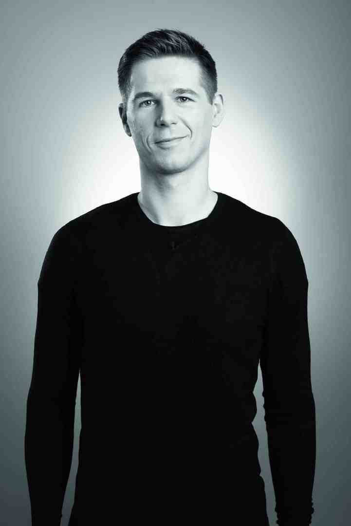 a man with a slight smile wearing a black longsleeve shirt standing against a light gray gradient background in a studio setting digital production A man with a slight smile wearing a black long-sleeve shirt, standing against a light gray gradient background in a studio setting.