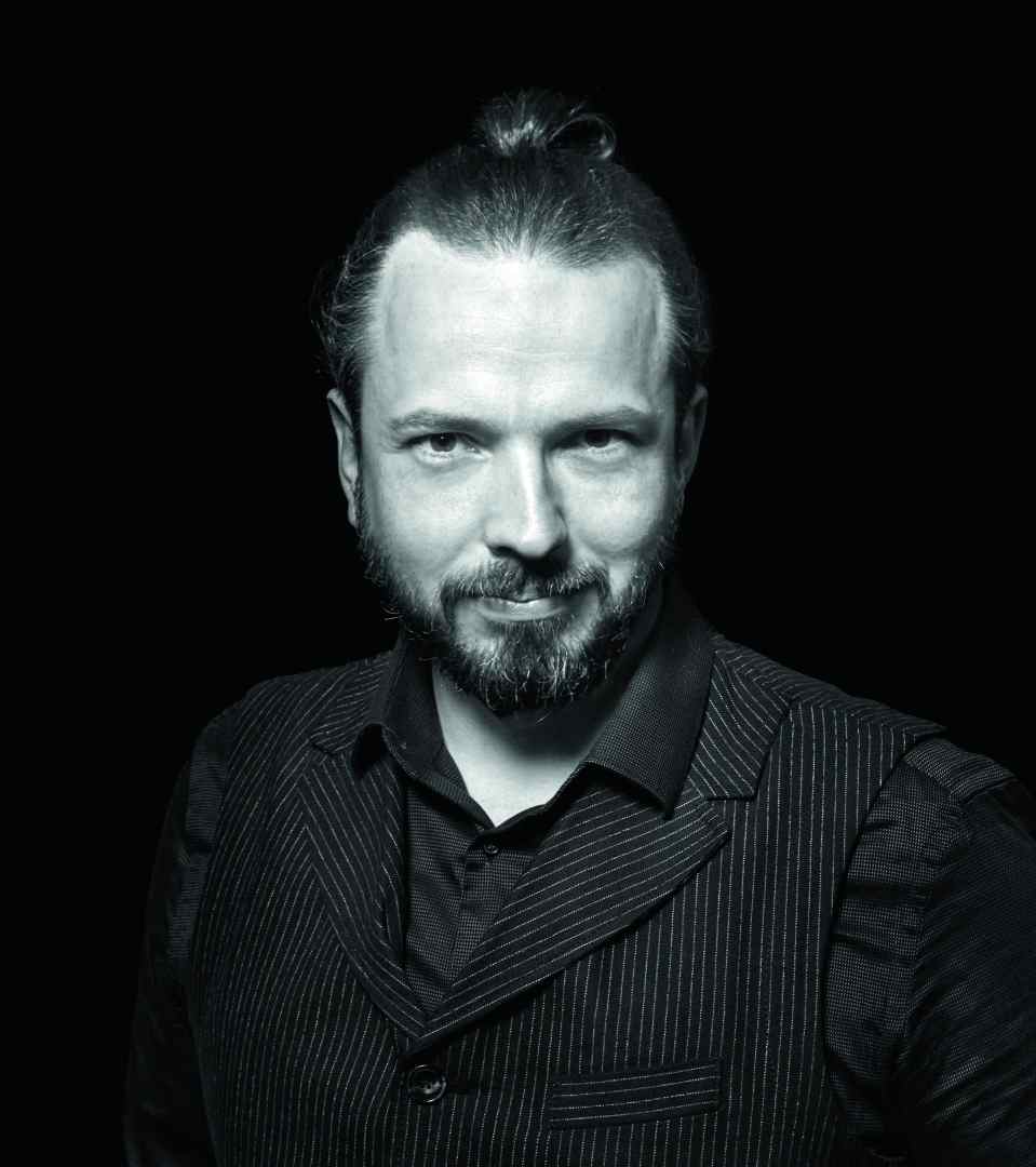a man with a beard and a top bun wearing a black pinstripe vest and shirt posing against a dark background with a confident expression digital production A man with a beard and a top bun wearing a black pinstripe vest and shirt, posing against a dark background with a confident expression.