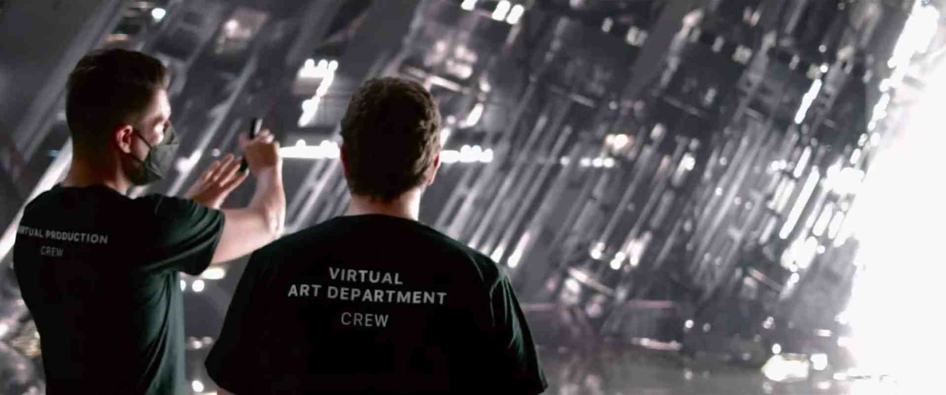 two crew members wearing black shirts stand in front of a reflective wall one taking a photo with a phone while the other gestures towards the installation highlighting their involvement in a virtual art production digital production Two crew members wearing black shirts stand in front of a reflective wall, one taking a photo with a phone while the other gestures towards the installation, highlighting their involvement in a virtual art production.