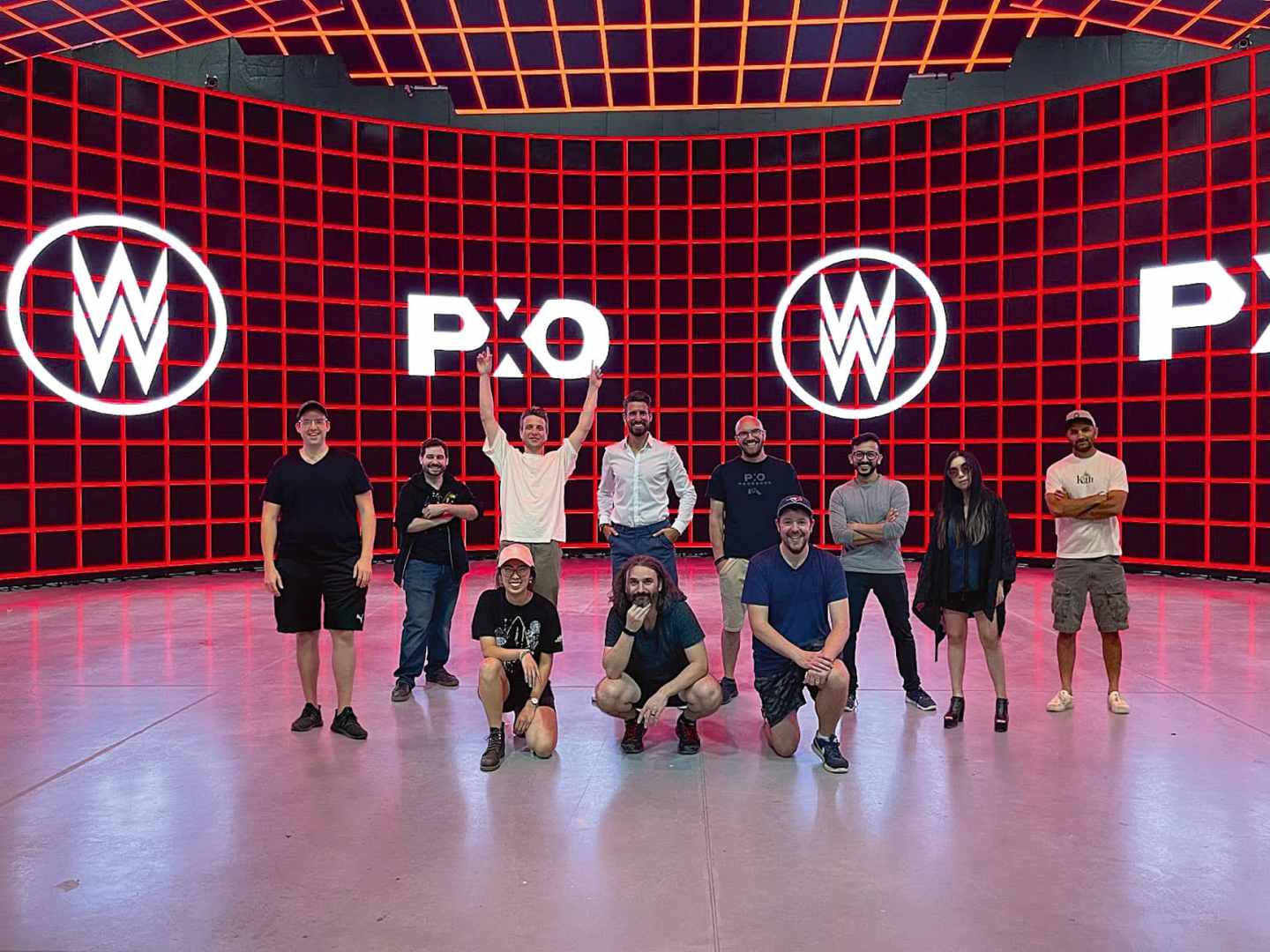 a group of ten people poses together in a vibrant performance space with large lighted screens displaying w and pxo logos in the background some individuals stand while others kneel showcasing smiles and celebratory gestures under colorful lighting digital production A group of ten people poses together in a vibrant performance space, with large lighted screens displaying