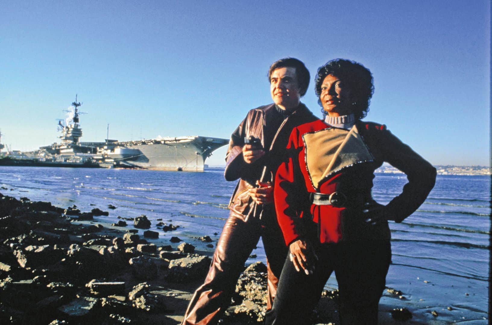 two individuals standing on rocky shore near a body of water with a large ship in the background one is wearing a red outfit with a decorative collar while the other wears a more casual outfit both looking towards the camera with confident expressions digital production Two individuals standing on rocky shore near a body of water, with a large ship in the background. One is wearing a red outfit with a decorative collar, while the other wears a more casual outfit, both looking towards the camera with confident expressions.