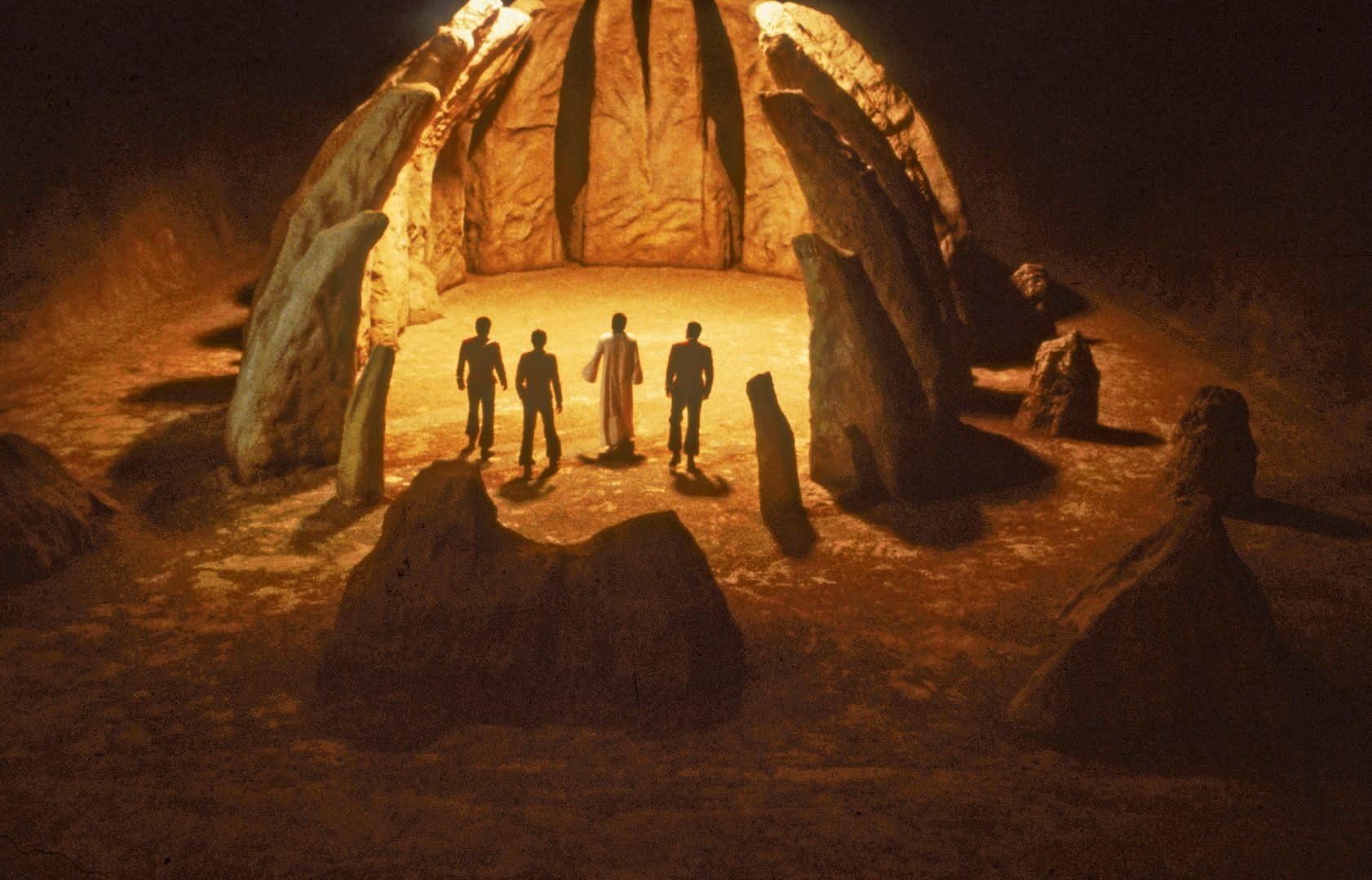 four silhouetted figures standing in a circular stone structure illuminated by warm light surrounded by tall rock formations in a dimly lit environment digital production Four silhouetted figures standing in a circular stone structure illuminated by warm light, surrounded by tall rock formations in a dimly lit environment.