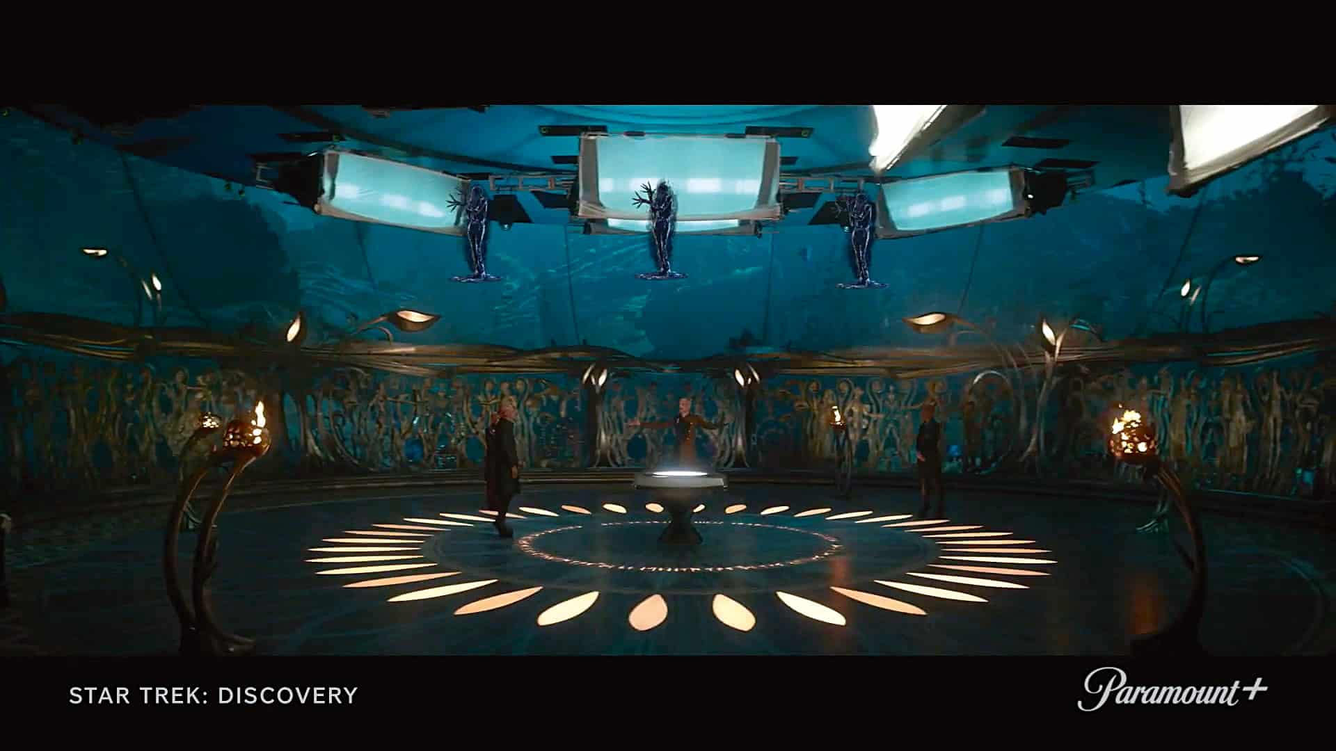 a futuristic room with a circular floor design featuring patterns of light tall blue walls with intricate artwork and holographic figures hovering overhead two figures stand in the room surrounded by glowing elements in a scifi environment digital production A futuristic room with a circular floor design featuring patterns of light. Tall, blue walls with intricate artwork and holographic figures hovering overhead. Two figures stand in the room, surrounded by glowing elements in a sci-fi environment.