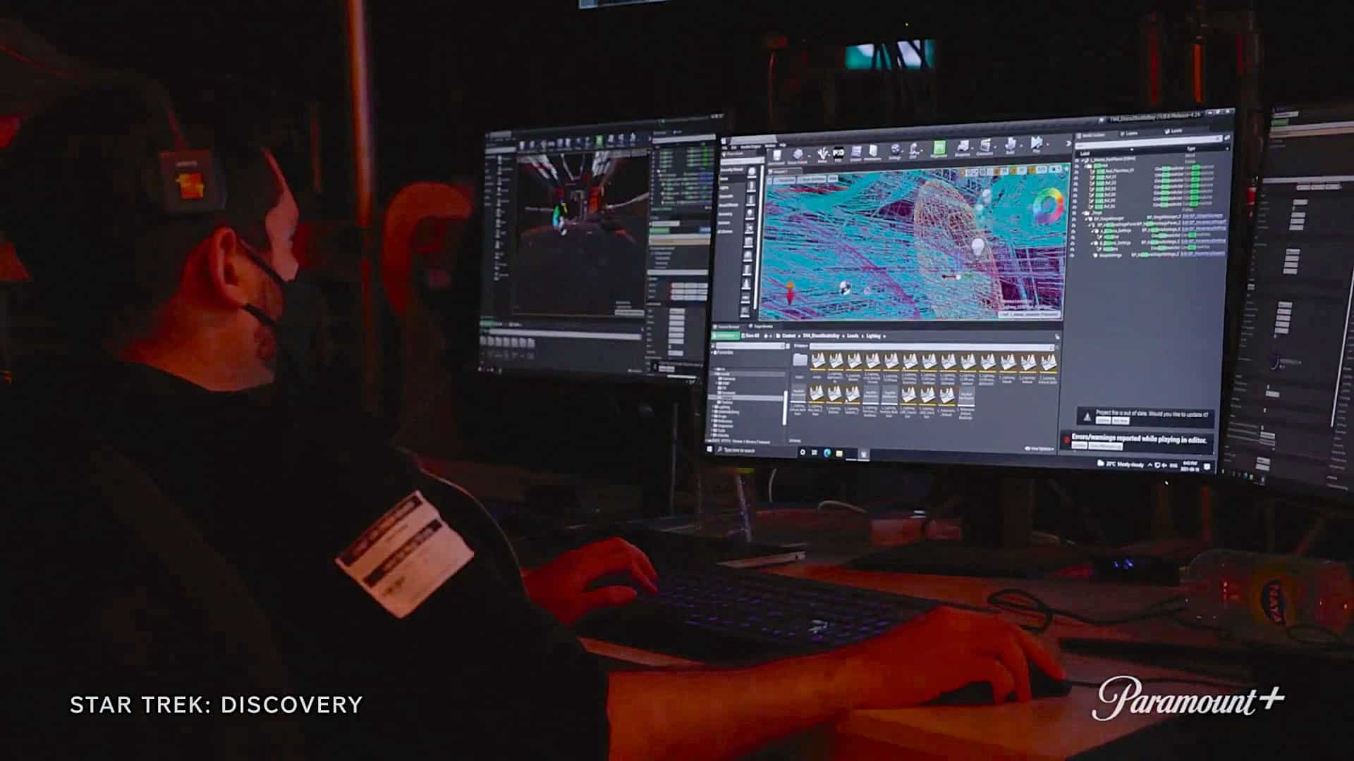 a person wearing a black shirt and mask sits in a dimly lit room focused on multiple computer monitors displaying graphics and animations related to a project the screens show colorful designs and data indicating a creative workspace digital production A person wearing a black shirt and mask sits in a dimly lit room, focused on multiple computer monitors displaying graphics and animations related to a project. The screens show colorful designs and data, indicating a creative workspace.