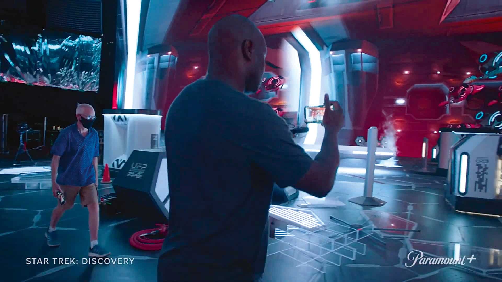 a man wearing a blue shirt and a mask takes a photo with a smartphone on a futuristic film set for star trek discovery another man in a black shirt walks in the background surrounded by advanced lighting and equipment digital production A man wearing a blue shirt and a mask takes a photo with a smartphone on a futuristic film set for 'Star Trek: Discovery.' Another man in a black shirt walks in the background, surrounded by advanced lighting and equipment.