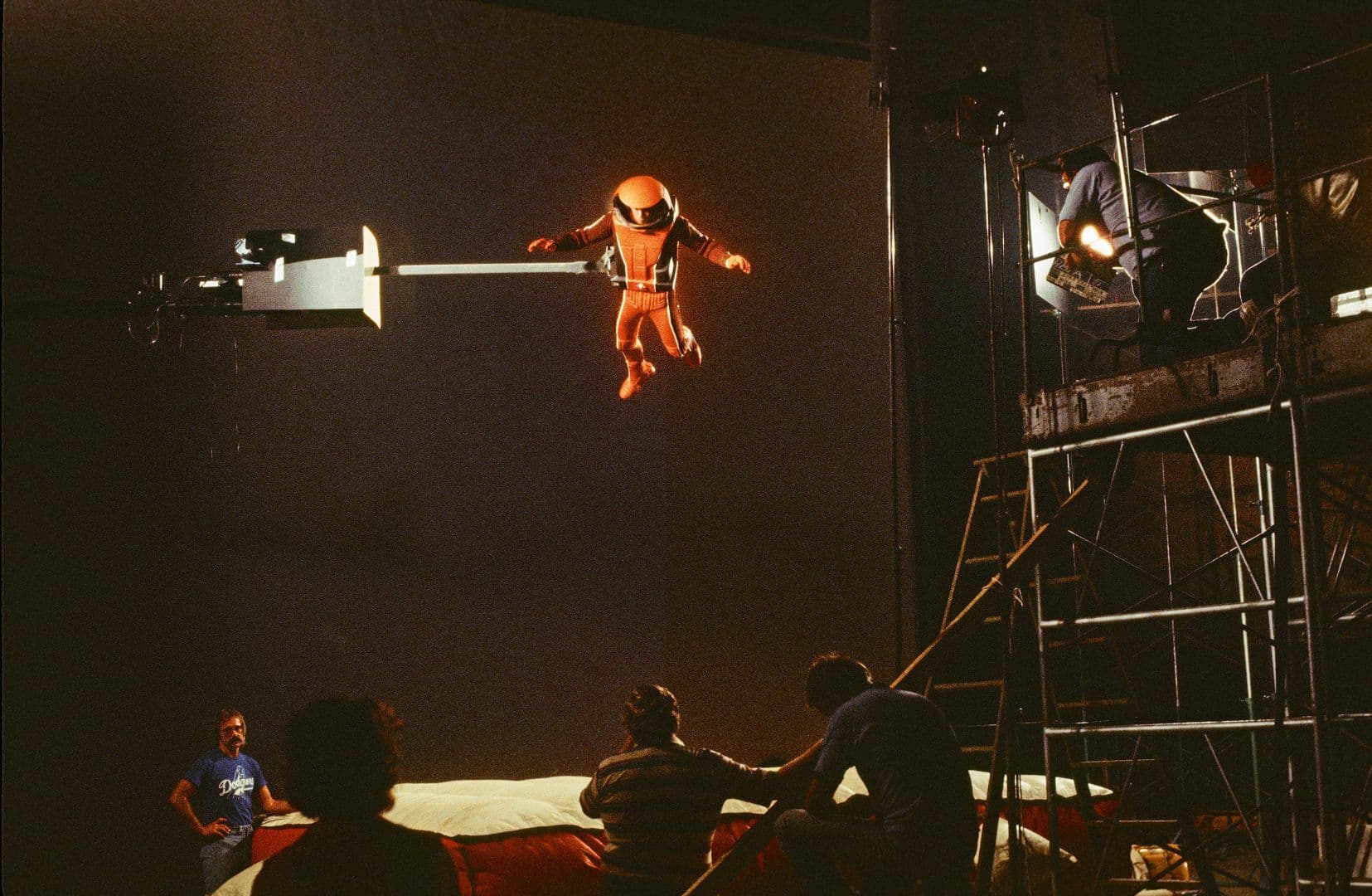 an astronaut in an orange suit is suspended above a padded surface on set with crew members gathered below scaffolding and lights reflect a film production environment digital production An astronaut in an orange suit is suspended above a padded surface on set, with crew members gathered below. Scaffolding and lights reflect a film production environment.