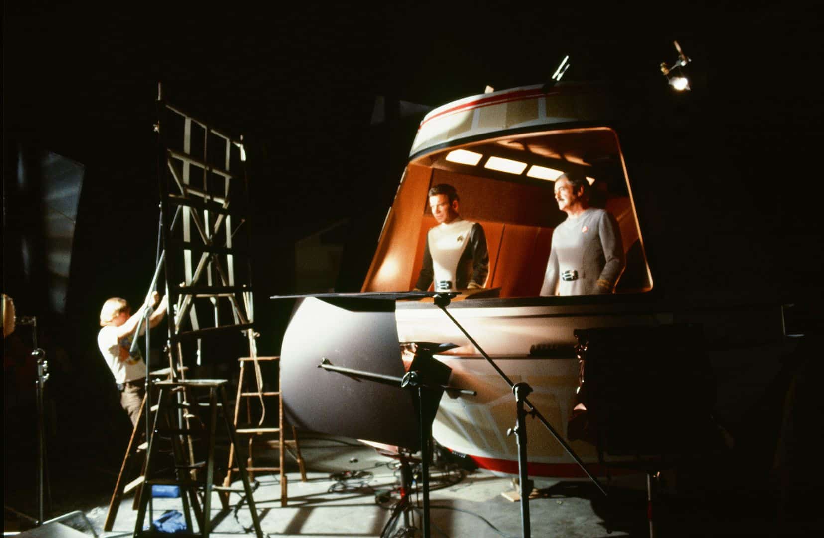 two actors in futuristic suits standing inside a spaceship set with warm lighting a crew member adjusts a ladder nearby surrounded by film equipment capturing an behindthescenes moment of a scifi production digital production Two actors in futuristic suits standing inside a spaceship set with warm lighting. A crew member adjusts a ladder nearby, surrounded by film equipment, capturing an behind-the-scenes moment of a sci-fi production.