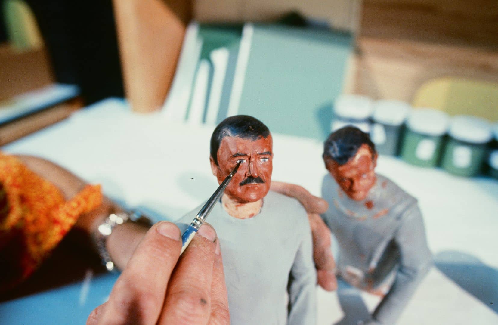 a closeup of a persons hand applying paint to the face of a small figurine with a mustache another unpainted figurine stands beside it on a table tools and paint containers are visible in the background suggesting a crafting or modeling environment digital production A close-up of a person's hand applying paint to the face of a small figurine with a mustache; another unpainted figurine stands beside it on a table. Tools and paint containers are visible in the background, suggesting a crafting or modeling environment.
