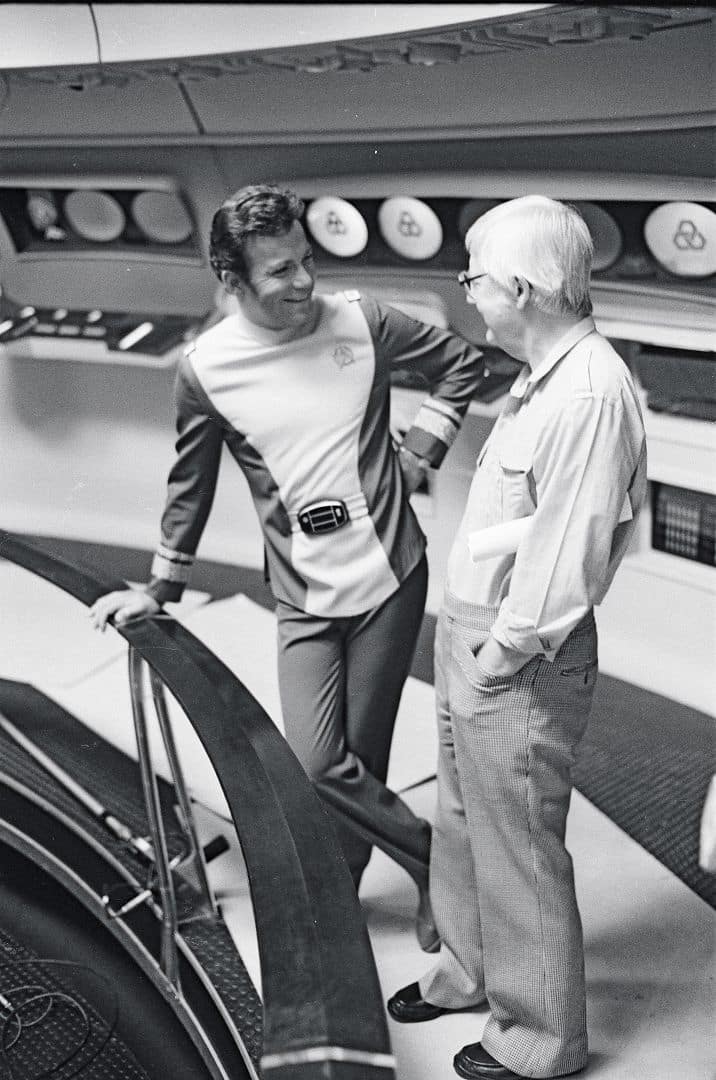a still image featuring two men engaged in conversation on a spaceship set one man wears a futuristic costume with a belt and a collar while the other wears glasses and casual attire both smiling as they chat the background shows circular elements typical of a scifi interior digital production A still image featuring two men engaged in conversation on a spaceship set. One man wears a futuristic costume with a belt and a collar, while the other wears glasses and casual attire, both smiling as they chat. The background shows circular elements typical of a sci-fi interior.