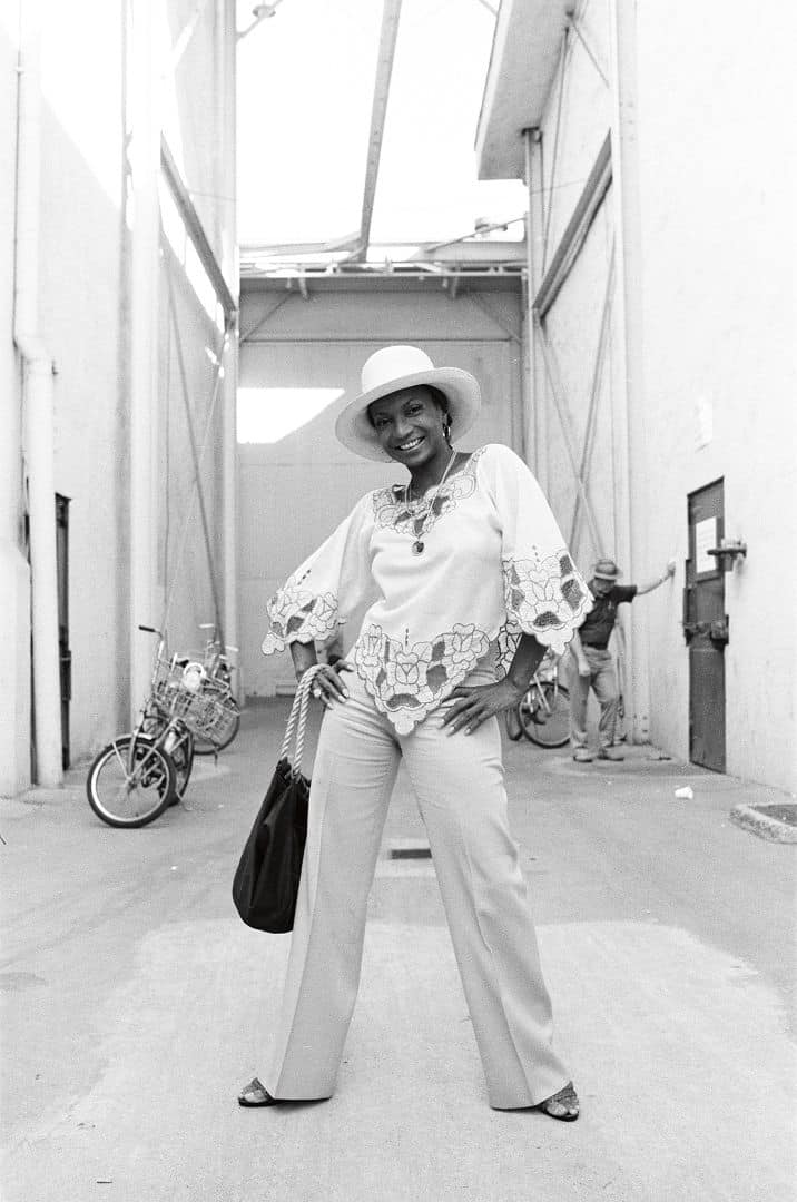 a woman in a stylish outfit featuring a patterned blouse and wideleg pants stands confidently in a narrow sunlit alley she wears a widebrimmed hat and holds a handbag with bicycles visible in the background digital production A woman in a stylish outfit featuring a patterned blouse and wide-leg pants stands confidently in a narrow, sunlit alley. She wears a wide-brimmed hat and holds a handbag, with bicycles visible in the background.