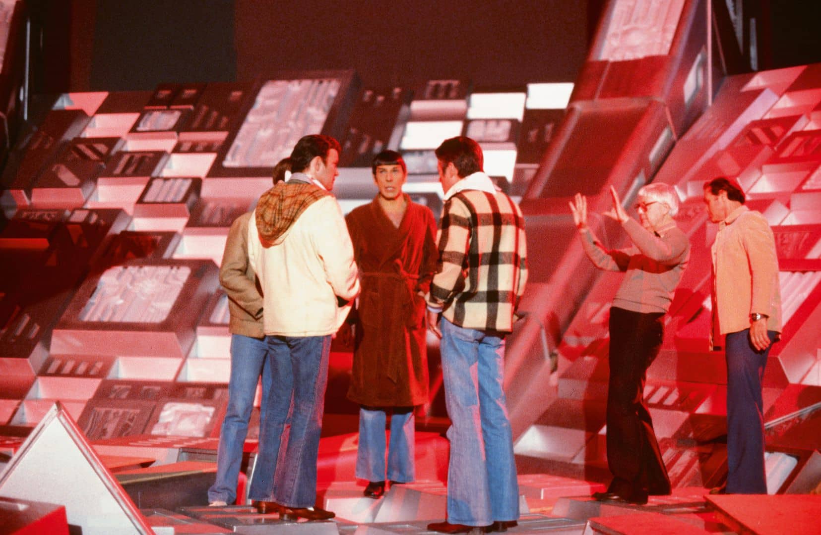 a group of six men on a brightly lit stage with a futuristic abstract background one man wears a robe while others are dressed in casual clothing engaged in conversation the setting features vibrant colors primarily red and silver creating an energetic atmosphere digital production A group of six men on a brightly lit stage with a futuristic, abstract background. One man wears a robe while others are dressed in casual clothing, engaged in conversation. The setting features vibrant colors, primarily red and silver, creating an energetic atmosphere.