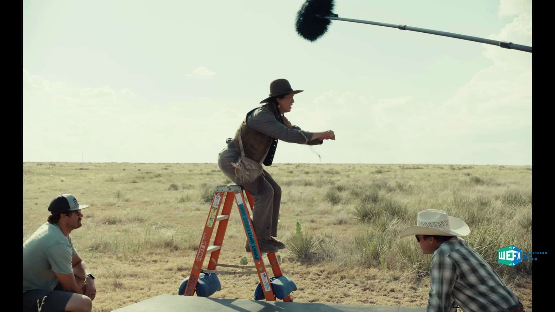 a person in a hat stands on a ladder while performing an action with two men sitting on the ground nearby likely observing a filming scene in a wideopen landscape under a cloudy sky digital production A person in a hat stands on a ladder while performing an action, with two men sitting on the ground nearby, likely observing a filming scene in a wide-open landscape under a cloudy sky.