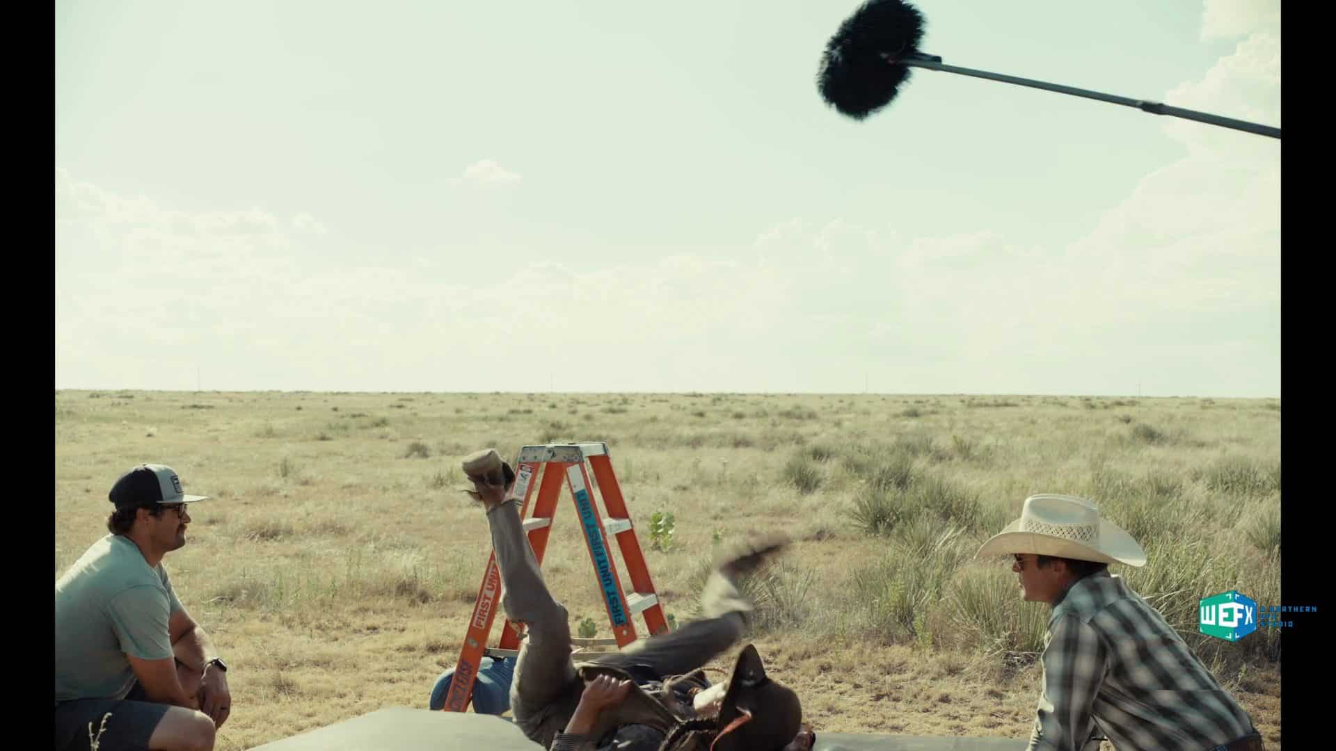 two men sit on a mat in a field observing a third man who has fallen over a microphone on a boom pole is being positioned above them a ladder is visible nearby in the grassy landscape under a cloudy sky digital production Two men sit on a mat in a field, observing a third man who has fallen over. A microphone on a boom pole is being positioned above them. A ladder is visible nearby in the grassy landscape under a cloudy sky.