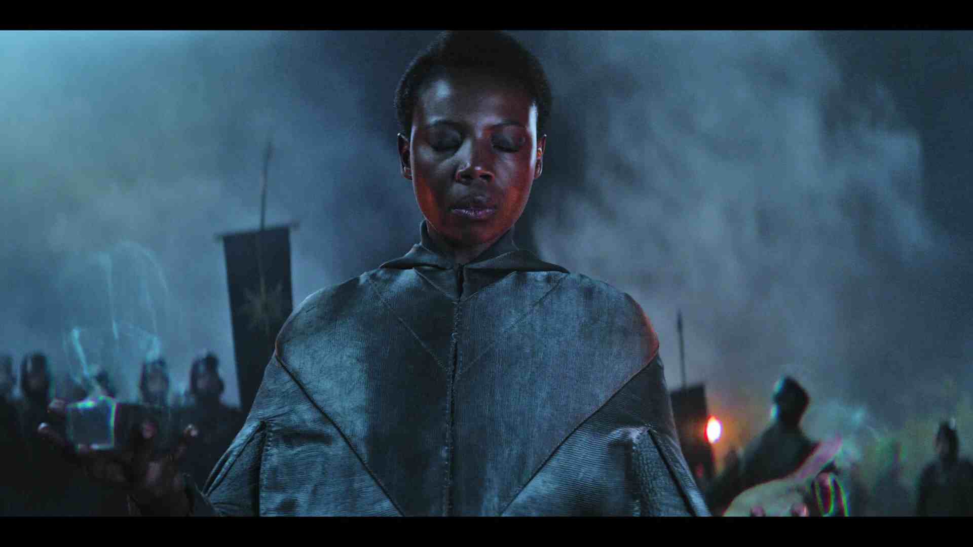 a woman wearing a dark textured cloak stands with an expression of concentration surrounded by a group of people in the background holding flags the scene is illuminated with a mystical glow against a smoky backdrop digital production A woman wearing a dark, textured cloak stands with an expression of concentration, surrounded by a group of people in the background holding flags. The scene is illuminated with a mystical glow against a smoky backdrop.