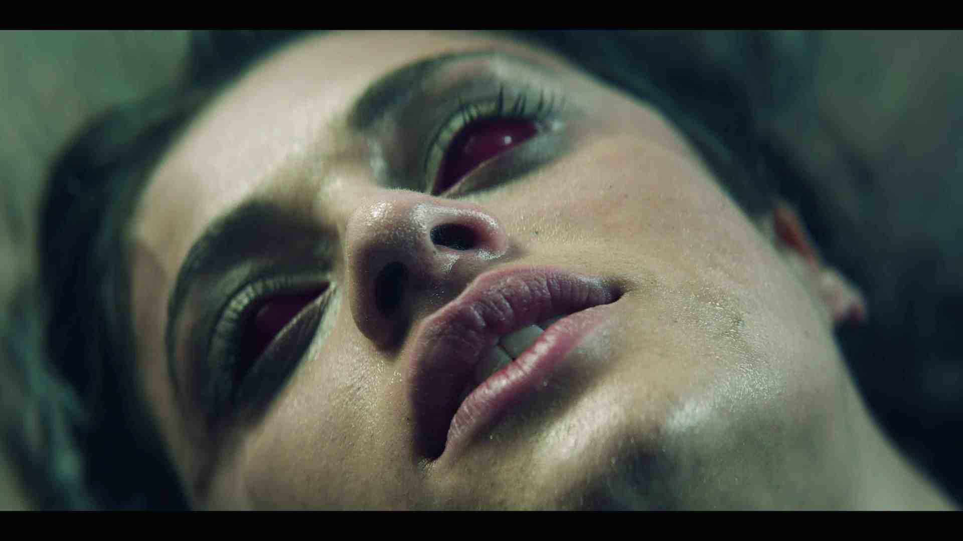 a closeup of a woman lying down her face glistening with moisture her eyes appear mostly red and her expression is neutral the background is blurred emphasizing her features digital production A close-up of a woman lying down with a distressed expression, featuring eerie red eyes and glistening skin.