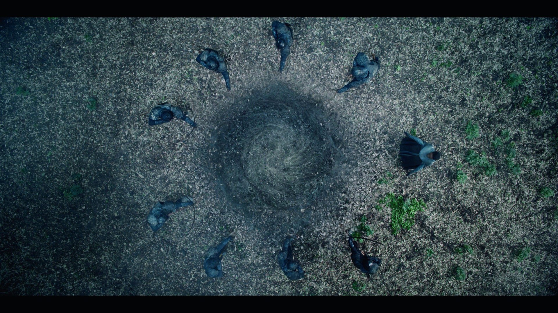 aerial view of a group of people dressed in dark clothing standing in a circle around a circular empty space on a forest floor covered with fallen leaves the center appears to be a disturbed area of soil digital production Aerial view of a group of people dressed in dark clothing standing in a circle around a circular, empty space on a forest floor covered with fallen leaves. The center appears to be a disturbed area of soil.