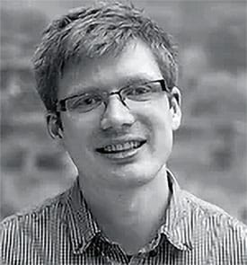 a closeup portrait of a young man with light brown hair and glasses smiling warmly at the camera he wears a collared shirt with thin stripes and the background is softly blurred focusing on his friendly expression digital production A close-up portrait of a young man with light brown hair and glasses, smiling warmly at the camera. He wears a collared shirt with thin stripes, and the background is softly blurred, focusing on his friendly expression.