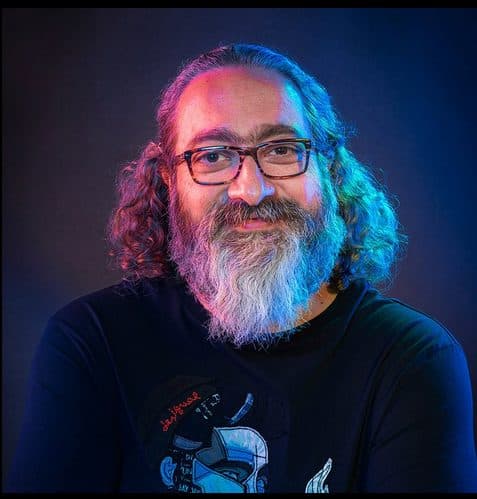 a man with curly gray hair and a beard wearing glasses and a black tshirt with a graphic design smiling at the camera against a dark background illuminated with blue and purple lighting digital production A man with curly gray hair and a beard, wearing glasses and a black t-shirt with a graphic design, smiling at the camera against a dark background illuminated with blue and purple lighting.