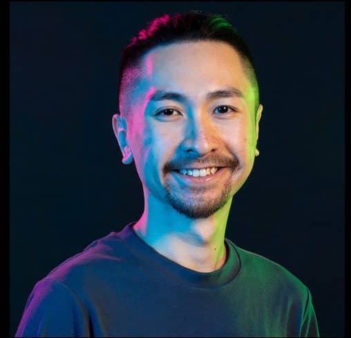 a smiling man with short hair and facial hair wearing a gray shirt is posing against a dark background illuminated with vibrant blue and green lighting digital production A smiling man with short hair and facial hair, wearing a gray shirt, is posing against a dark background illuminated with vibrant blue and green lighting.