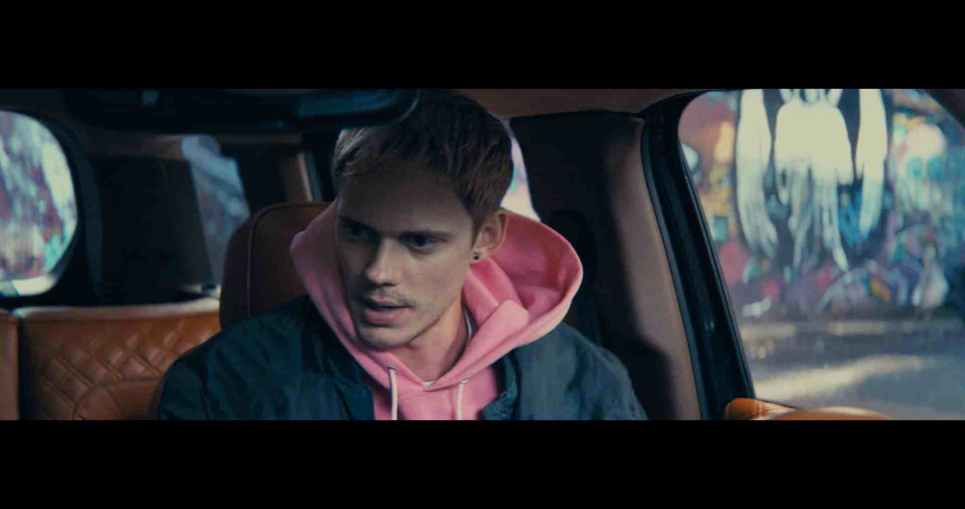 a young man with light hair and a serious expression wearing a pink hoodie under a dark jacket sits inside a vehicle the background features colorful street art creating an urban atmosphere digital production A young man with light hair and a serious expression, wearing a pink hoodie under a dark jacket, sits inside a vehicle. The background features colorful street art, creating an urban atmosphere.