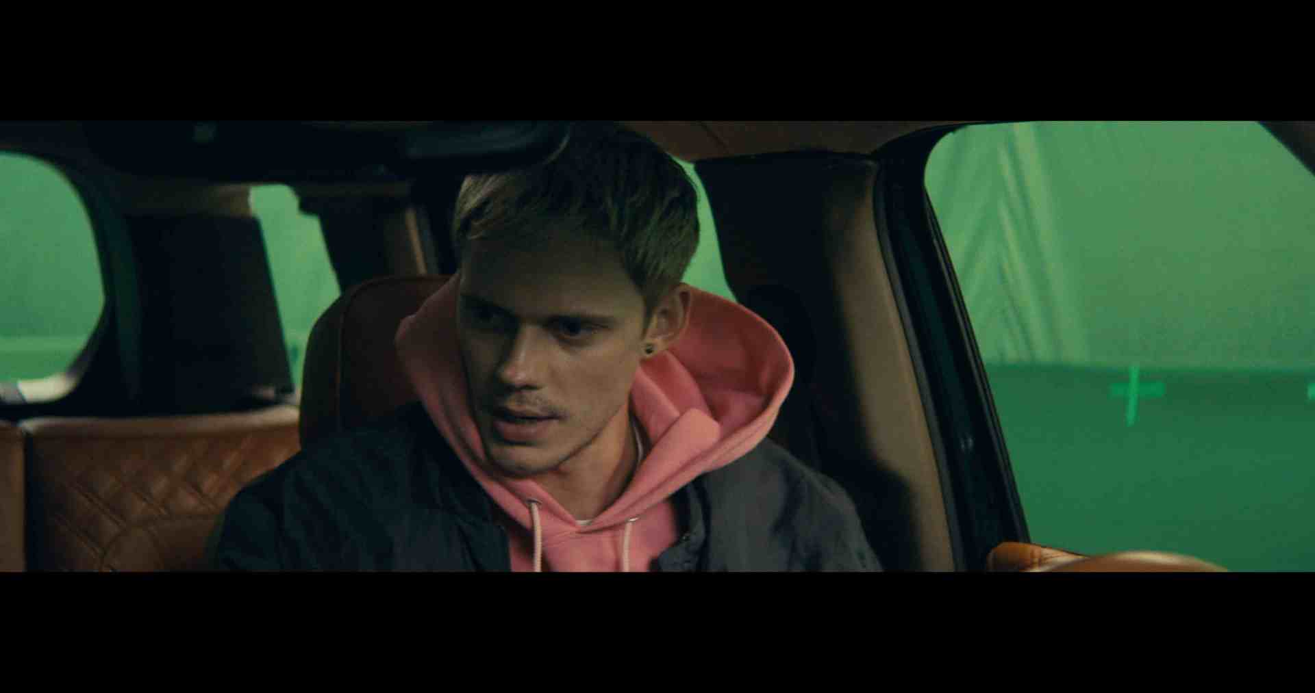 a young man with short blonde hair wearing a pink hoodie and a dark jacket sits in the front seat of a car the background features green screens indicating a film or photo shoot setting digital production A young man with short blonde hair wearing a pink hoodie and a dark jacket sits in the front seat of a car. The background features green screens, indicating a film or photo shoot setting.