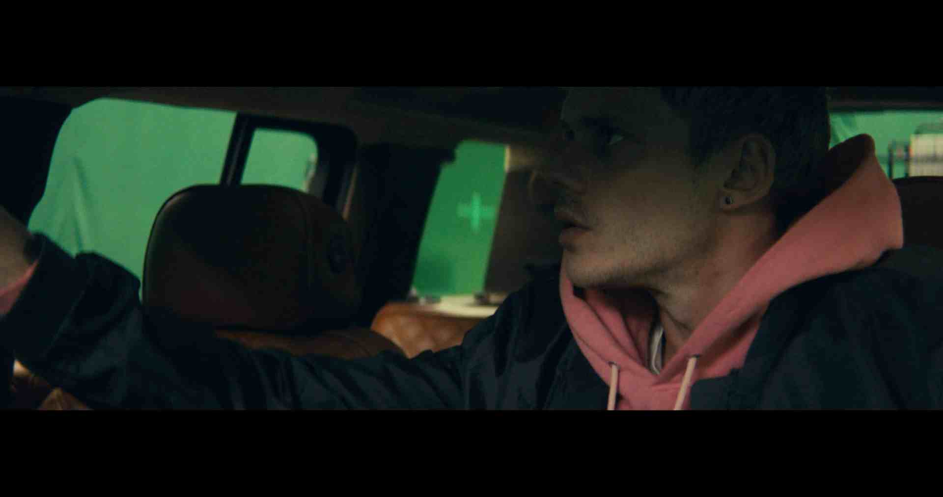a man with short hair wearing a pink hoodie and a dark jacket sits in the backseat of a car looking intently to the side the background features green screen elements suggesting a film or video production setting digital production A character inside a vehicle looking towards the dashboard, with green screen elements in the background.