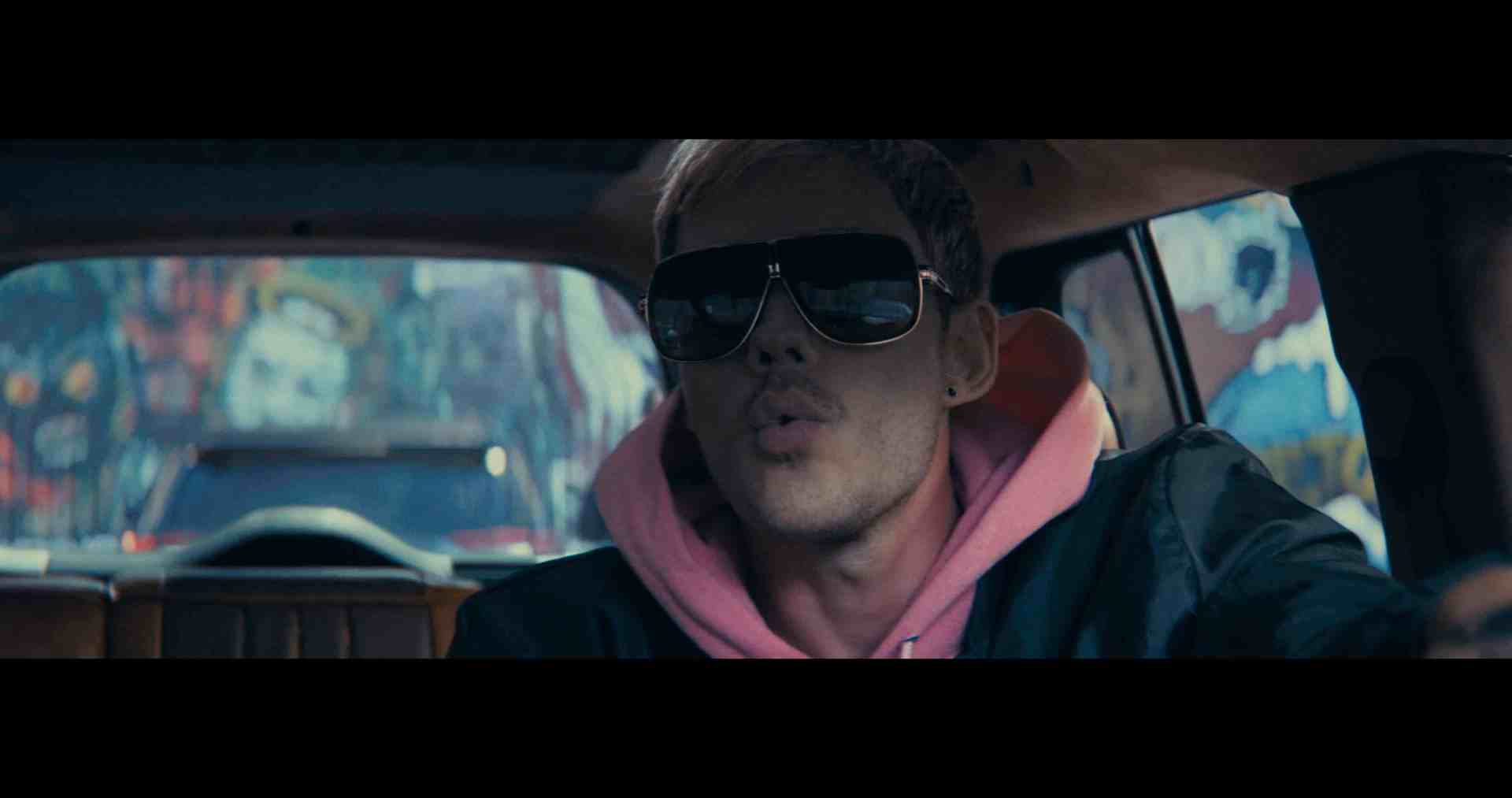 a man wearing oversized sunglasses and a pink hoodie sits in the drivers seat of a car with a blurred street art background visible through the window he appears to be focused and animated digital production A person wearing stylish sunglasses and a pink hoodie sits in a car, exhaling while facing forward. The background features colorful graffiti art.