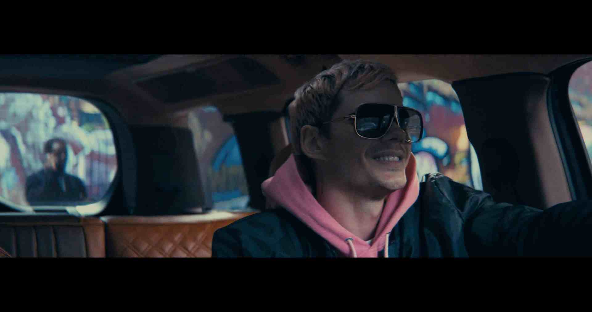 a person with blonde hair wearing sunglasses and a pink hoodie is driving a vehicle the interior features a brown quilted design and vibrant graffiti art is visible in the background through the windows digital production A person with blonde hair wearing sunglasses and a pink hoodie is driving a vehicle. The interior features a brown quilted design, and vibrant graffiti art is visible in the background through the windows.