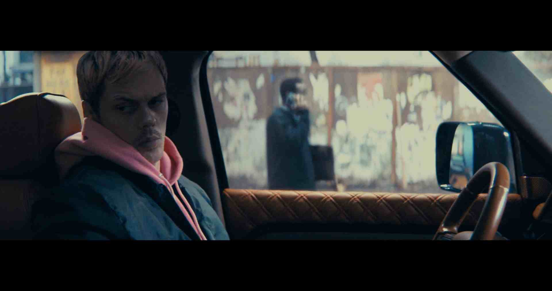 a young man with short bleached hair sits in the drivers seat of a luxury suv wearing a pink hoodie under a dark jacket in the background a man in dark clothes stands against a graffiticovered wall holding a phone digital production A man with short blonde hair, wearing a pink hoodie and black jacket, looks pensively out the window of a car, while a blurred figure stands in the background talking on a phone.