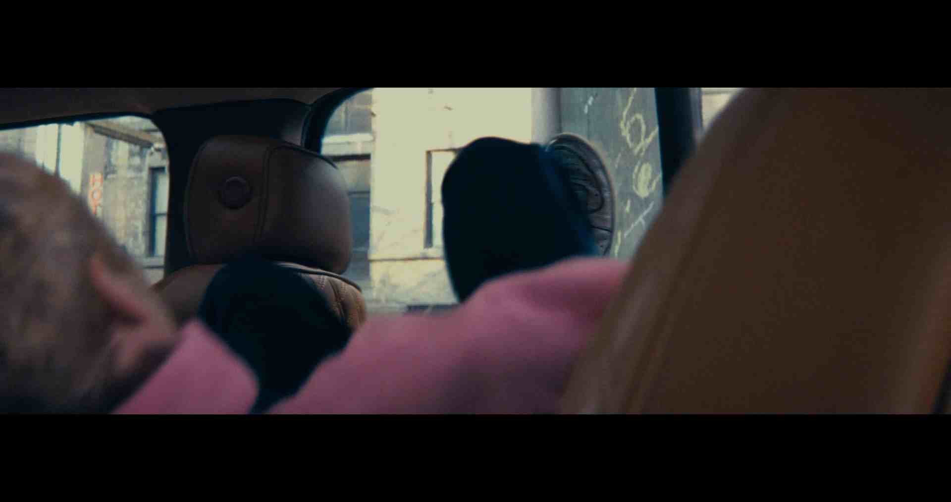 inside a vehicle two individuals with dark clothing relax in their seats one with a hoodie while the other leans back a blurred urban scene is visible through the open window showcasing the exterior of a building digital production Interior of a car with a focus on the back seat, showing a foot resting on the seat and part of a person's head visible in the foreground.