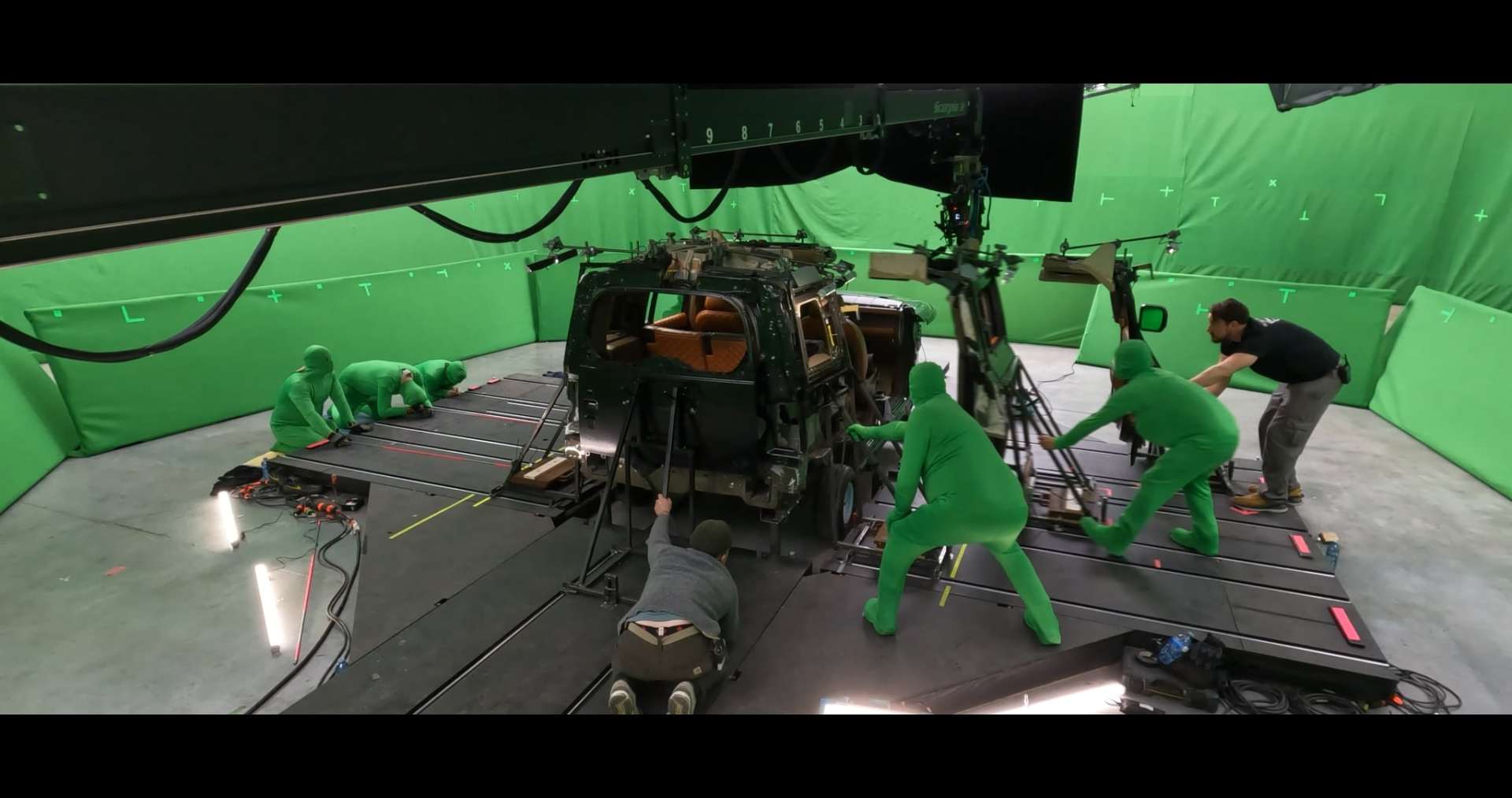 a team of crew members in green motion capture suits working around a partially built vehicle on a film set with a greenscreen backdrop preparing for special effects and filming digital production A VFX team in green suits working on a car set surrounded by green screens, with equipment and lighting in a studio.