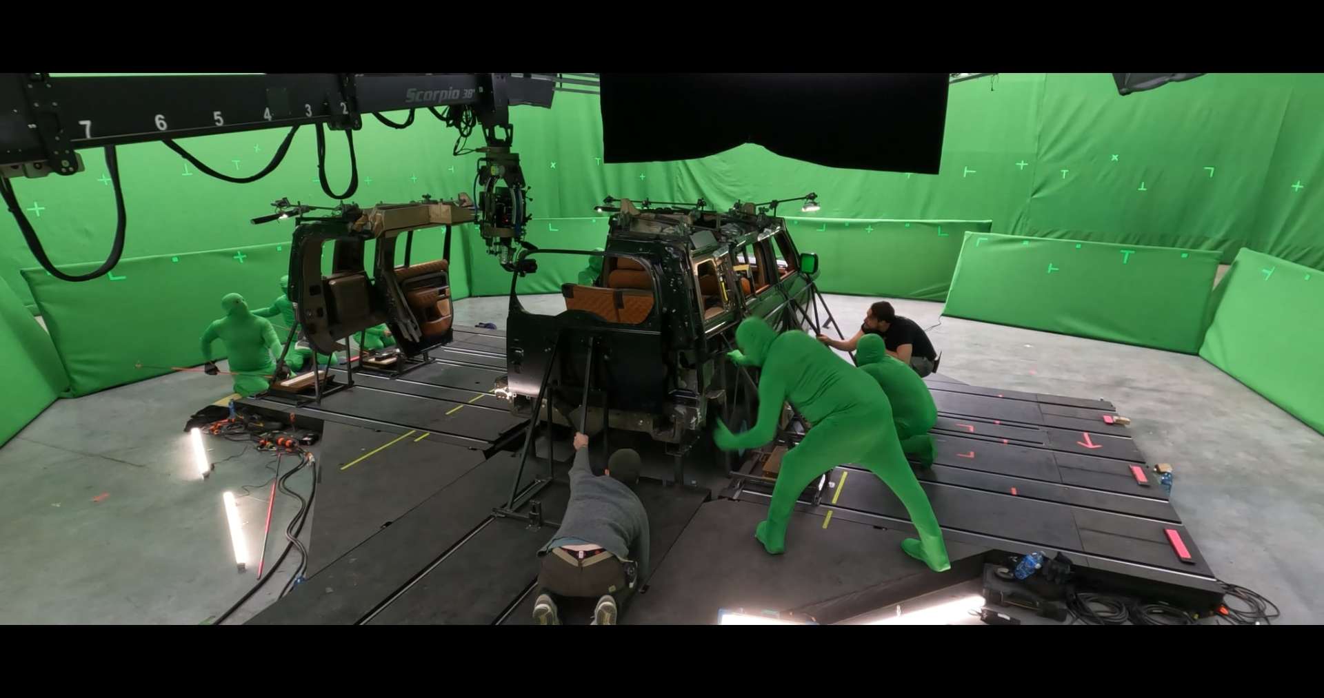 a film set featuring two vehicle mockups surrounded by crew members in green screen suits equipment and cables are visible on the floor with crew members adjusting the vehicles and preparing for a shoot digital production A film crew working on a car set surrounded by green screens. Crew members in green bodysuits are adjusting the vehicle, while others are focused on setups.