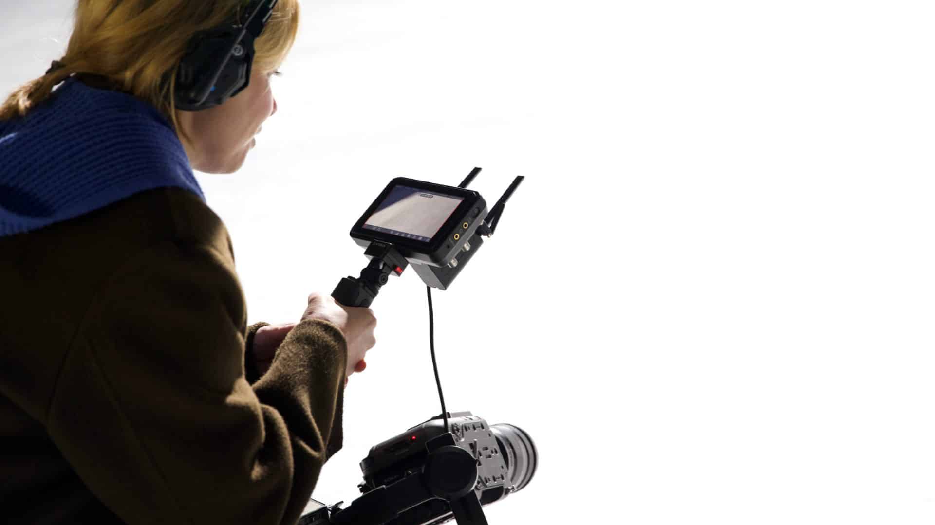 a woman wearing headphones operates a camera with a monitor mounted on top focusing intently on the shot she has short blonde hair and wears a dark coat with a blue collar soft lighting highlights her concentration digital production A woman wearing headphones operates a camera with a monitor mounted on top, focusing intently on the shot. She has short blonde hair and wears a dark coat with a blue collar. Soft lighting highlights her concentration.