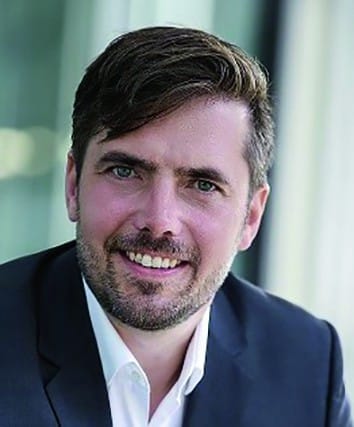 a man with dark hair and a beard smiles warmly at the camera wearing a black suit jacket over a white shirt soft natural light highlights his facial features against a blurred modern background digital production A man with dark hair and a beard smiles warmly at the camera, wearing a black suit jacket over a white shirt. Soft natural light highlights his facial features against a blurred modern background.