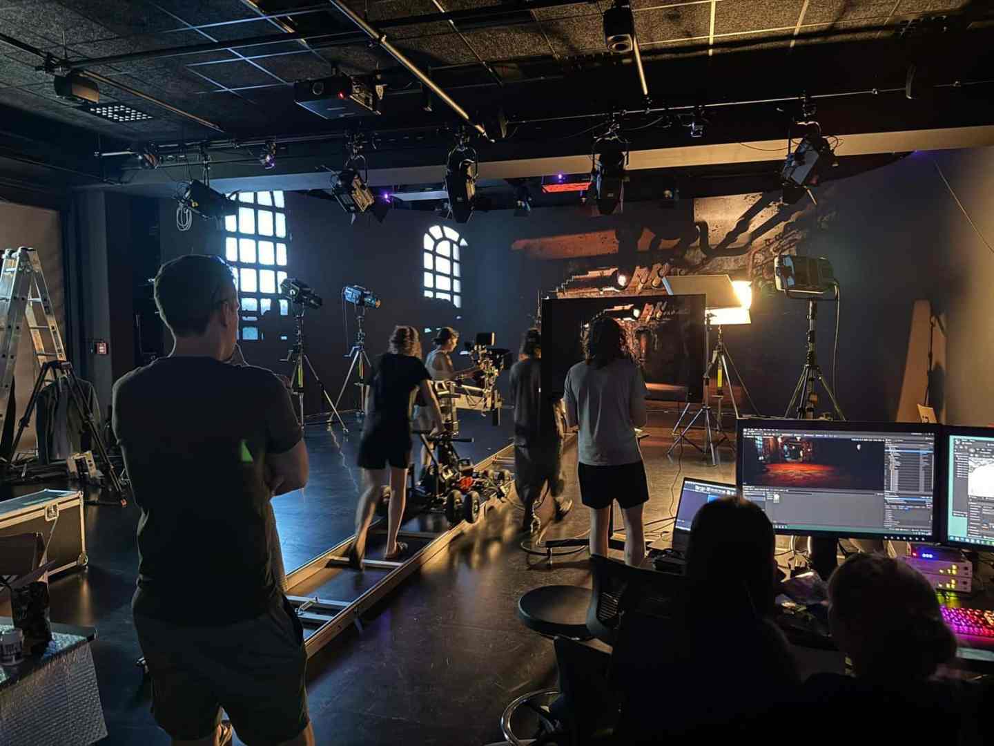 a film studio setup with multiple crew members working bright lights and cameras are positioned around a dark room two people are operating computer equipment while others adjust lighting and camera angles digital production A film studio setup with multiple crew members working. Bright lights and cameras are positioned around a dark room. Two people are operating computer equipment, while others adjust lighting and camera angles.