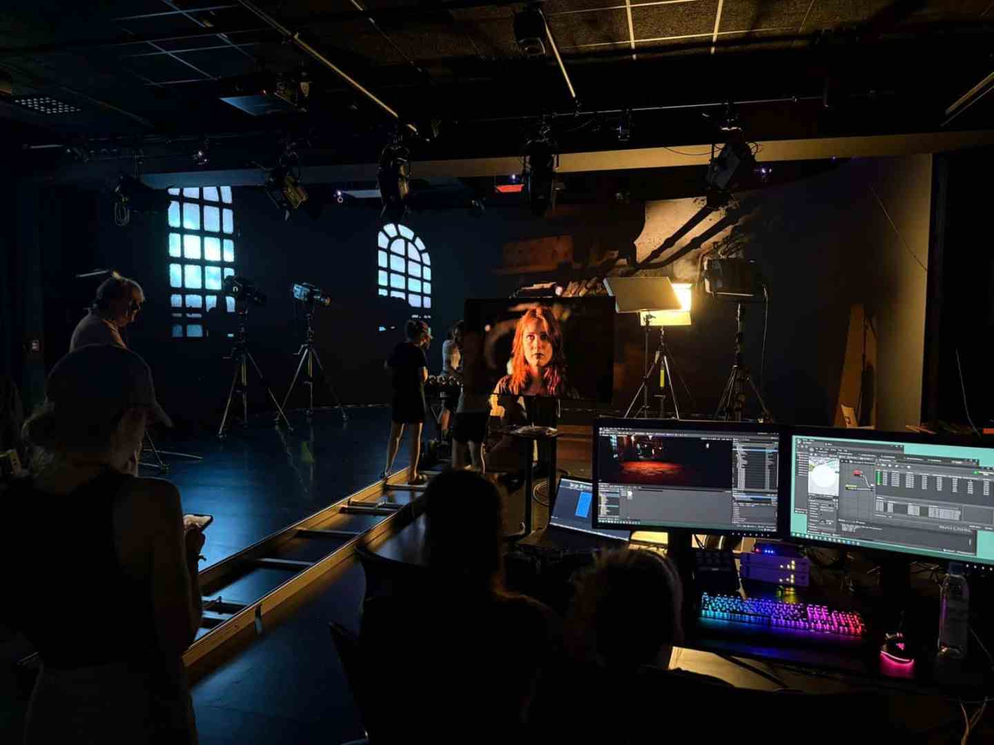 a film studio scene with crew members surrounding a bright spotlight focused on a female subject cameras are set up with various lighting and monitors display editing software the space is dimly lit with colorful window patterns in the background digital production A film studio scene with crew members surrounding a bright spotlight focused on a female subject. Cameras are set up with various lighting, and monitors display editing software. The space is dimly lit with colorful window patterns in the background.