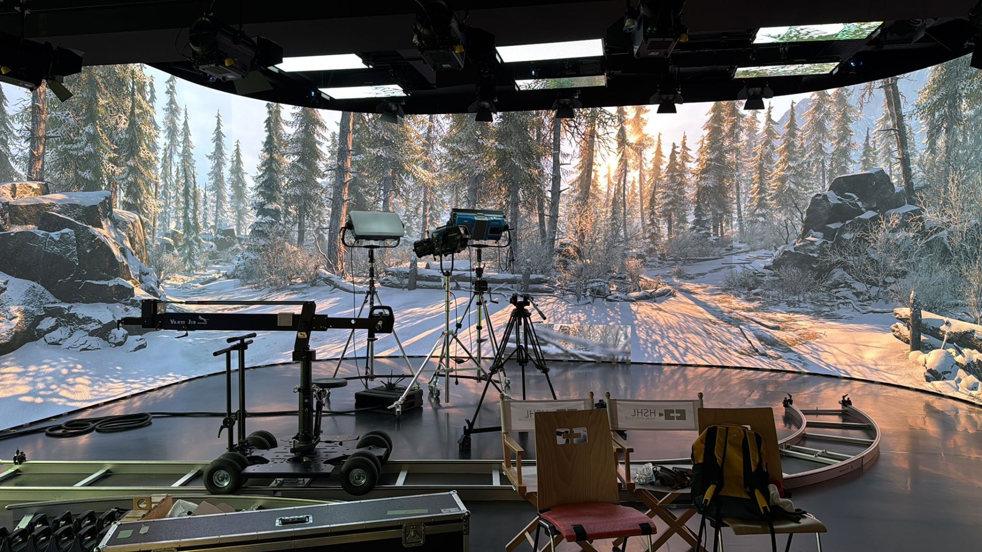 a film studio set showcasing a winter forest scene with snowy ground and tall trees in the background equipment like cameras and chairs are visible emphasizing the production environment digital production A film studio set showcasing a winter forest scene with snowy ground and tall trees in the background. Equipment like cameras and chairs are visible, emphasizing the production environment.